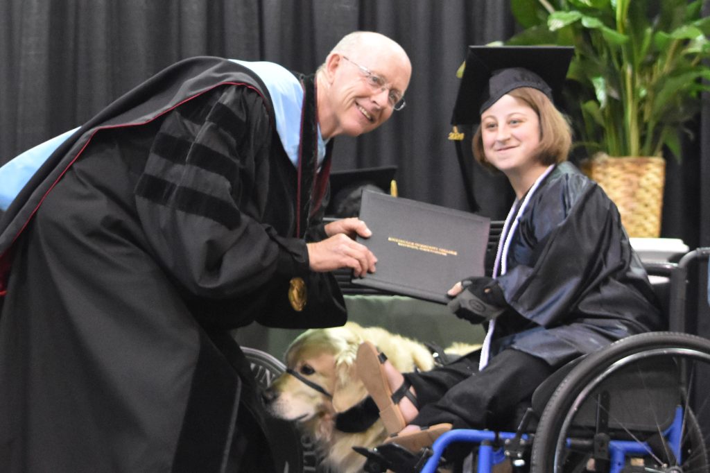 Female student in a wheelchair with a guide dog accepts her degree from the college president