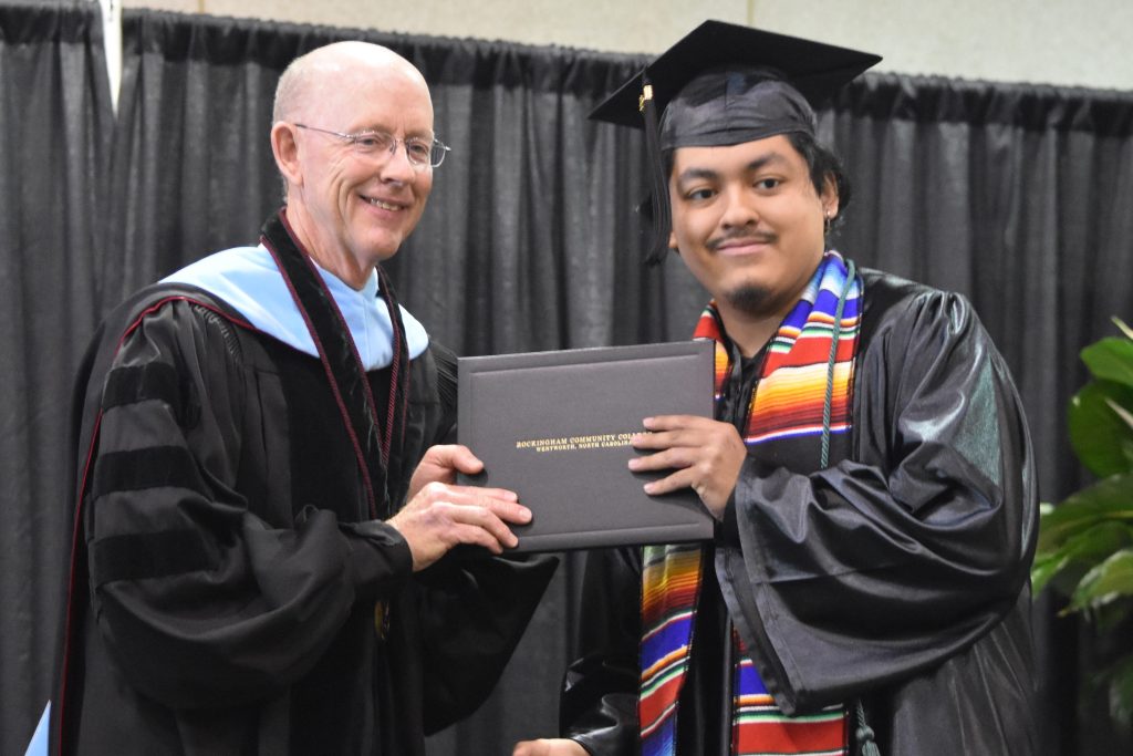 Male Hispanic student in rainbow sash accepts his degree from the college president