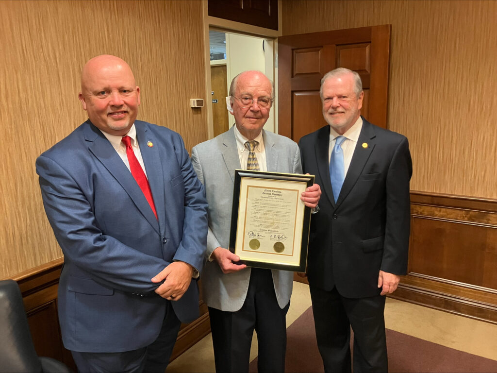 Three men in suits smile, with the middle one holding a framed certificate.