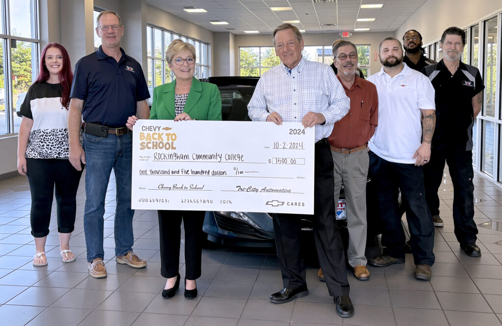 A group of people gathered in a car dealership's showroom holding a gigantic $1,500 check from Tri-City Automotive to Rockingham Community College.