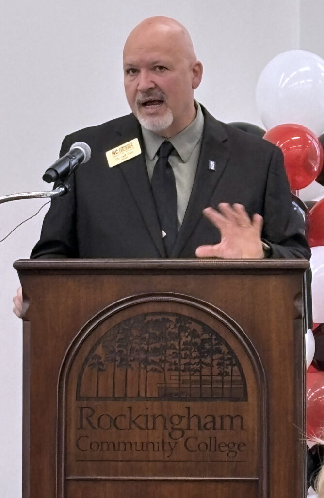 Photo of a man speaking at a podium carved with the words Rockingham Community College and some trees. Balloons are in the background.