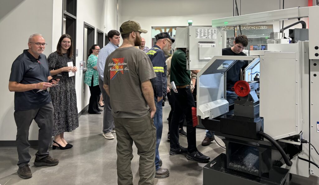 Several adults gather around machinery as one student demonstrates how it works.