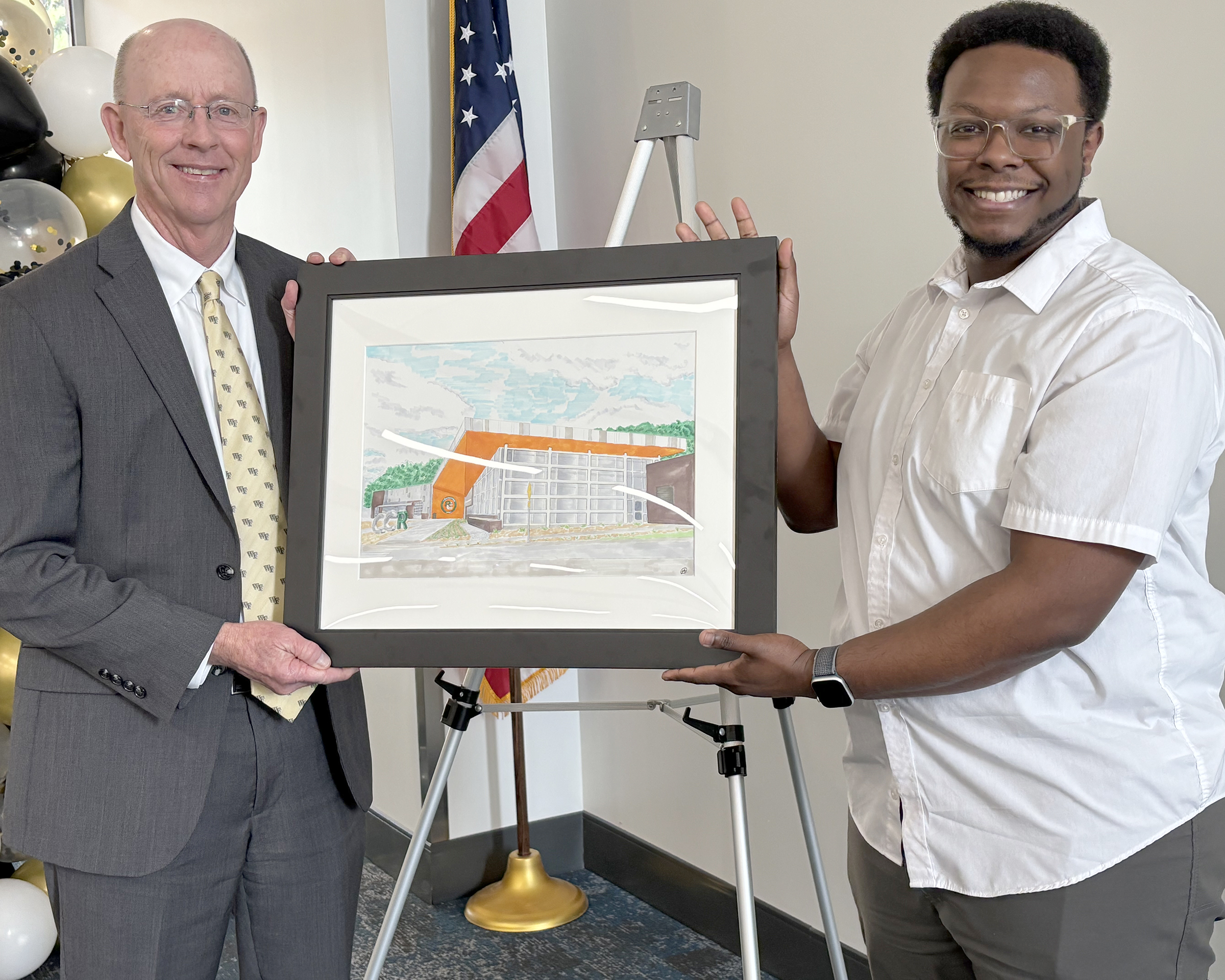 Two men hold framed artwork depicting a building.