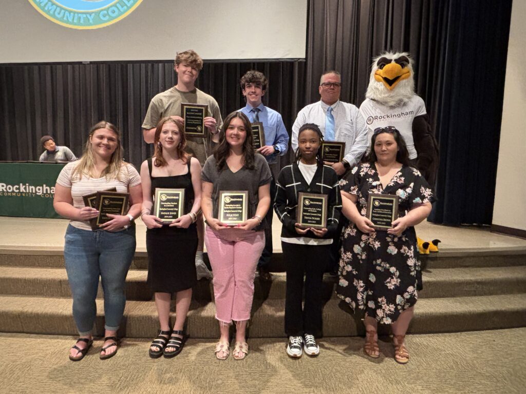 Eight college students display plaques on a stage with an eagle mascot.