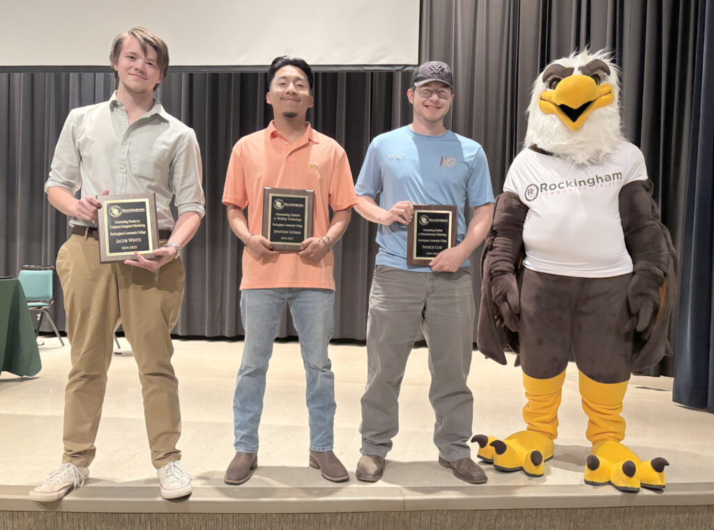 Three college students display plaques on a stage with an eagle mascot.