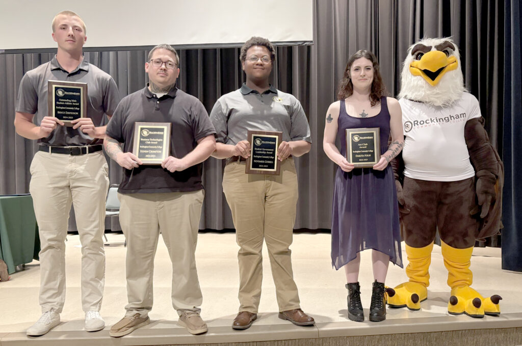Four college students display plaques on a stage with an eagle mascot.