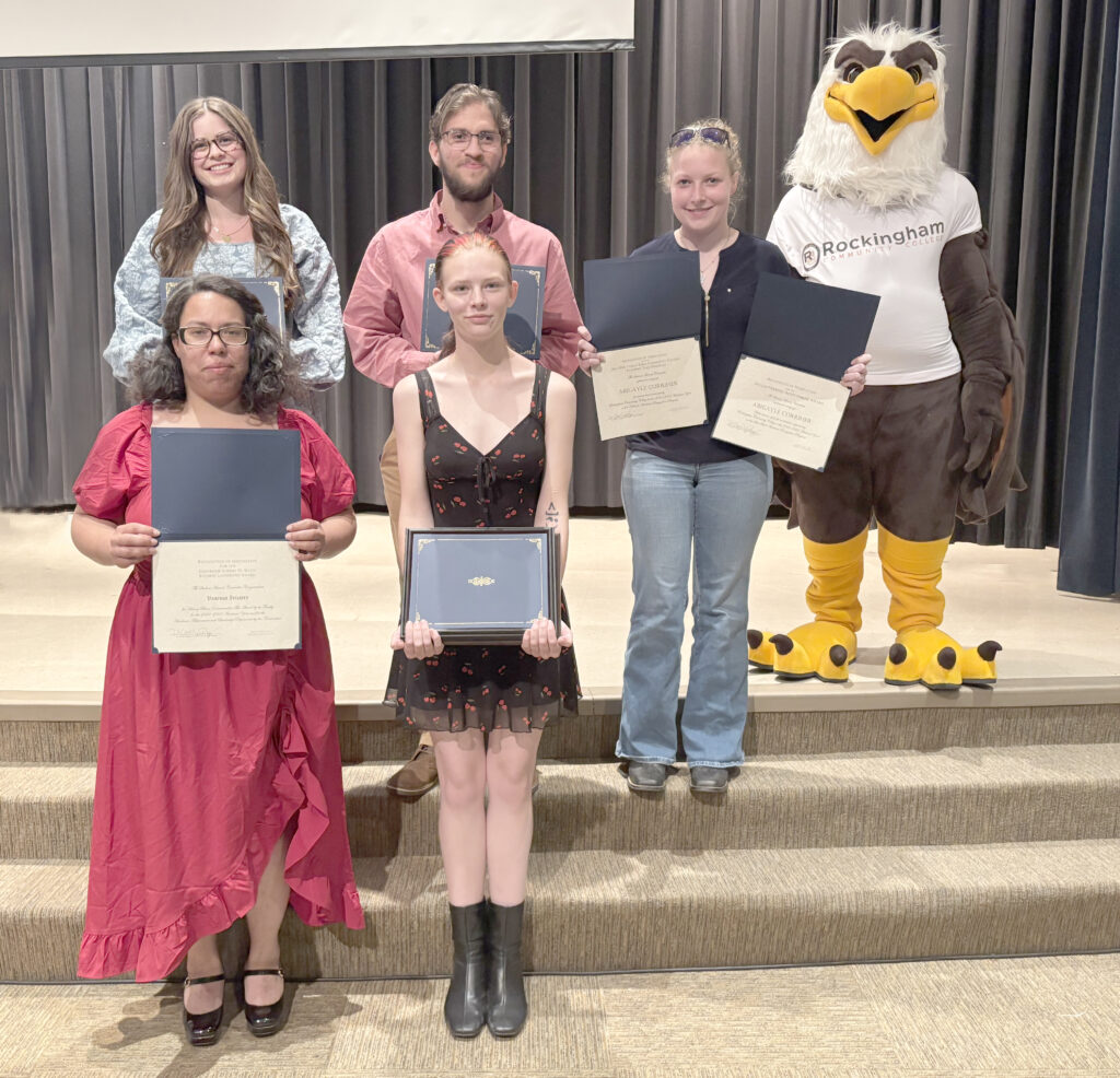Five college students display plaques on a stage with an eagle mascot.