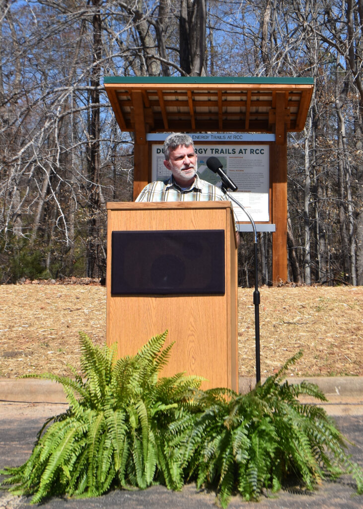 A man talks at a podium in front of a sign that says, Duke Energy Trails at RCC.