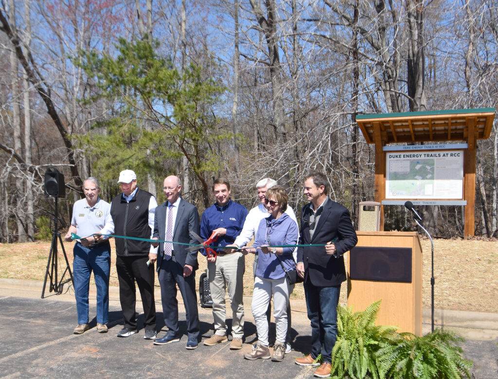 A group of adults hold a green ribbon, with one in the center about to cut the ribbon with a large pair of scissors.