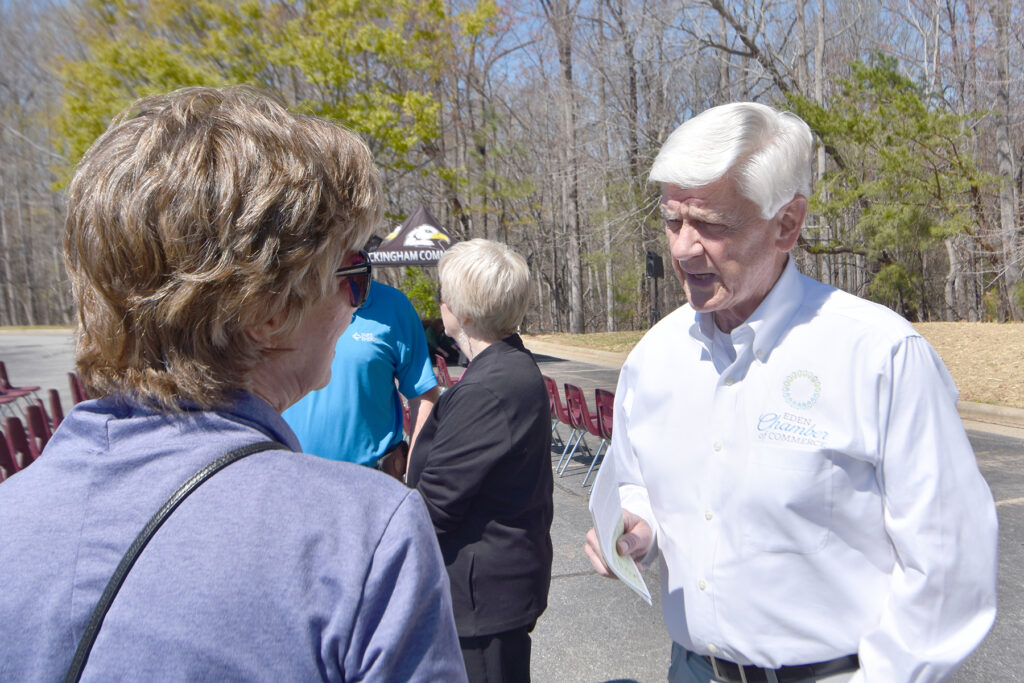 A woman and man talk in a parking lot, surrounded by trees and rows of chairs in the background.