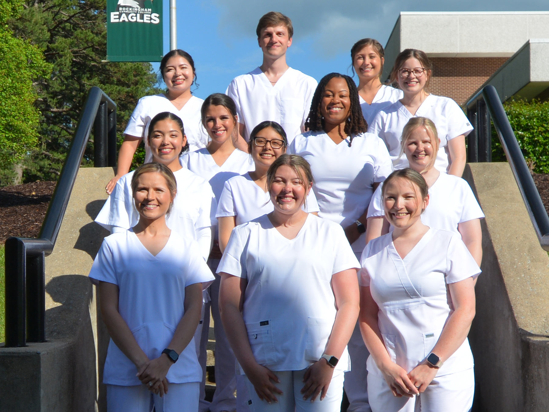 Eleven young women and one young man, all in white scrubs, smile on a staircase, with a banner in the background that says Rockingham Community College Eagles.