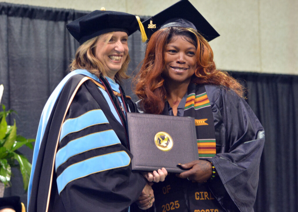 Two women shake hands and hold a diploma on stage.