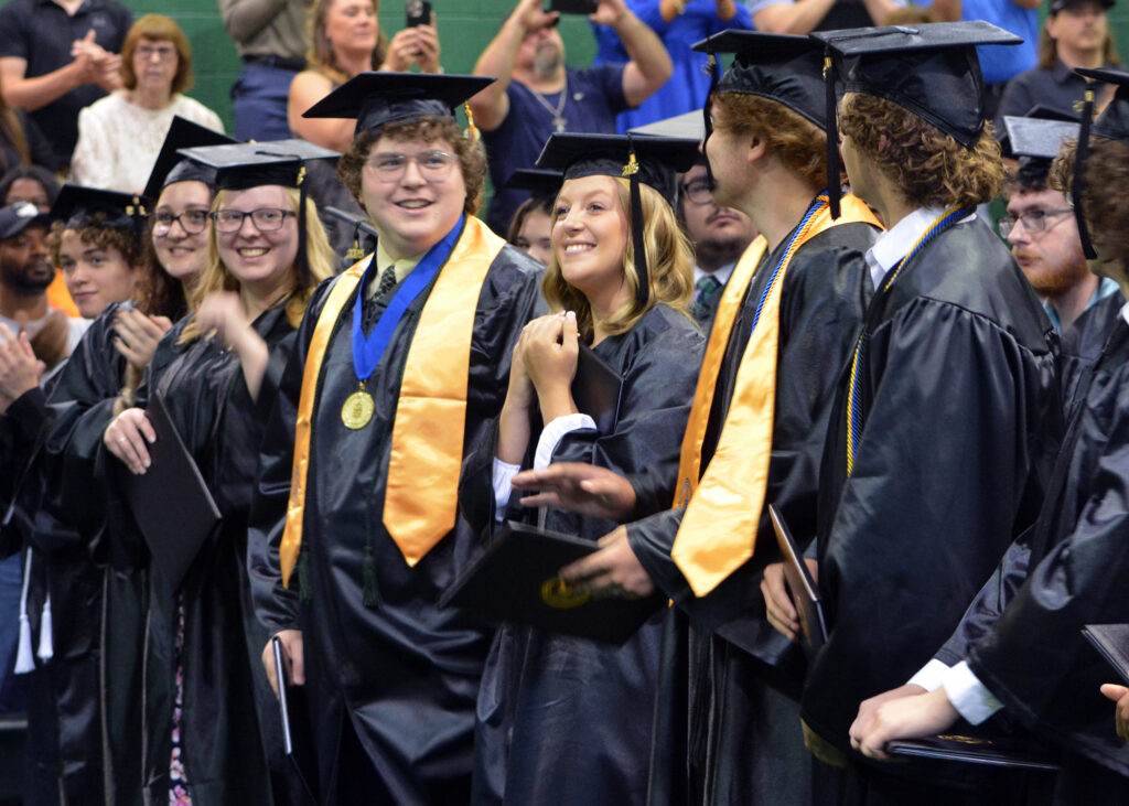A female graduate grips her diploma and smiles as male and female graduates smile, clap, and look at each other from the front row during the graduation ceremony after turning their tassels.