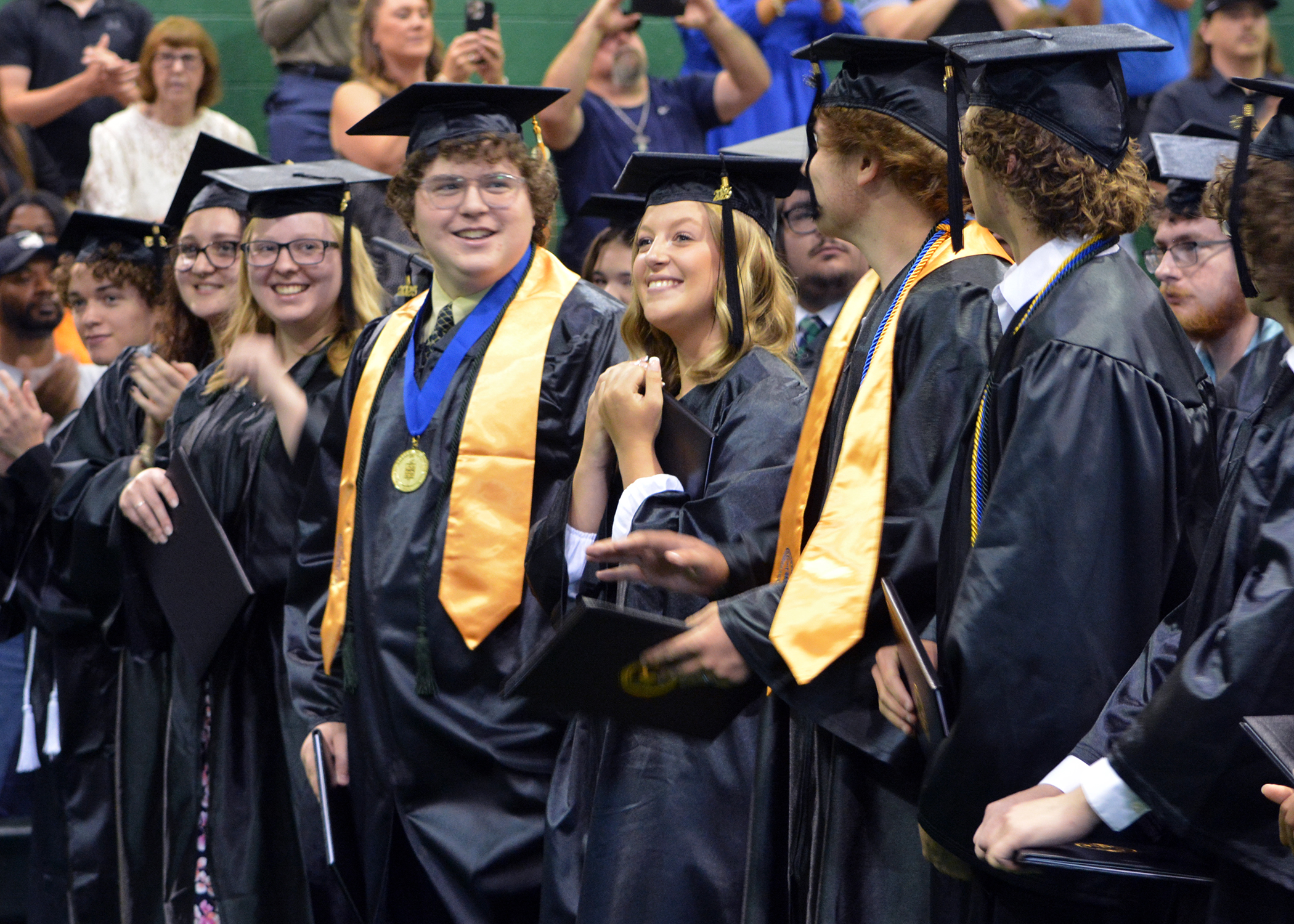 A female graduate grips her diploma and smiles as male and female graduates smile, clap, and look at each other from the front row during the graduation ceremony after turning their tassels.