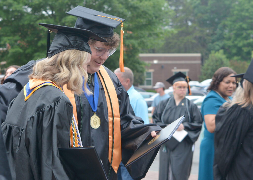 A male graduate smiles as he looks at the cover of his diploma with a female graduate, both in their caps and gowns.