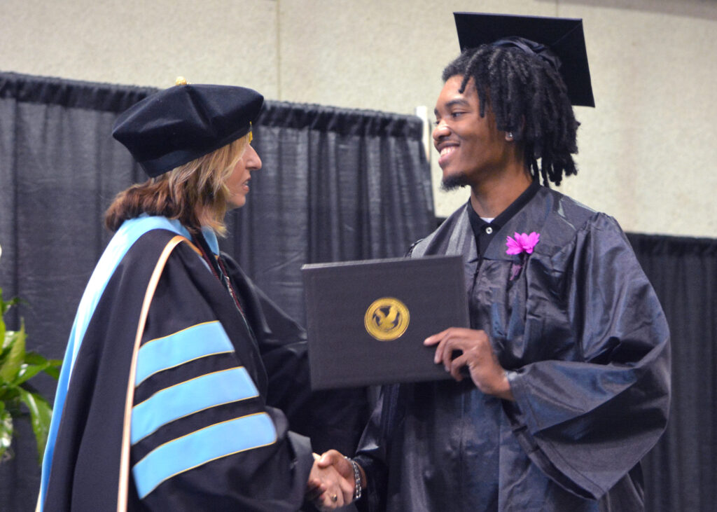 A female shakes hands with a male graduate on stage as she hands him his diploma.