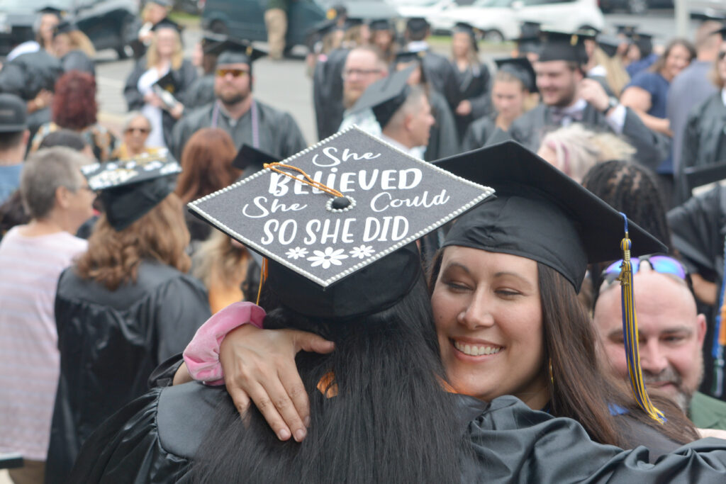 A female faculty member hugs a graduate who is wearing a cap with the words she believed she could so she did, with many other graduates in the background.