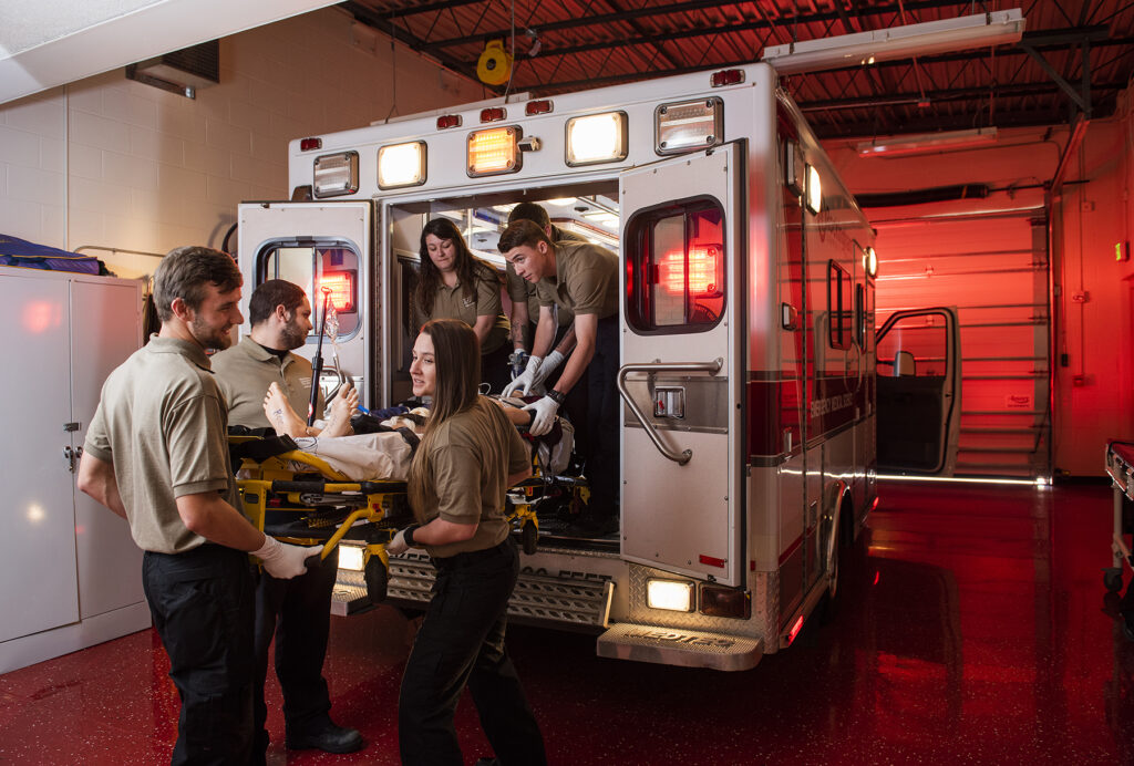 EMT students unload a stretcher with a mannequin from the back of an ambulance.