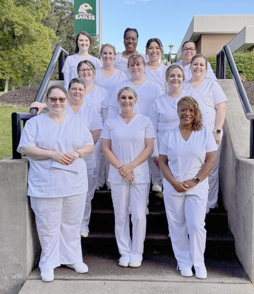 A group of young women in white scrubs smile on a staircase, with a banner in the background that says Rockingham Community College Eagles.