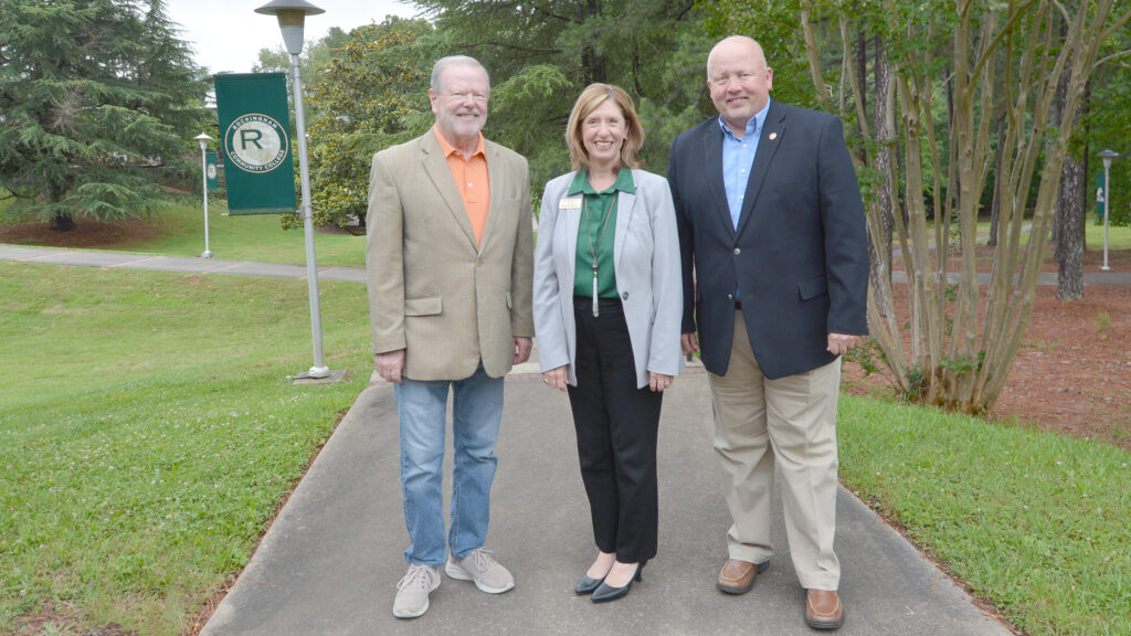 Two men and a woman smile on a sidewalk outside that has lamp posts with Rockingham Community College banners hanging from them.