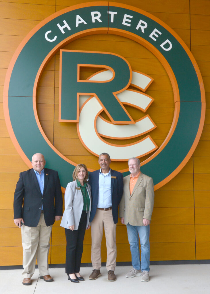 Three men and a woman smile in front of a giant Rockingham Community College logo.