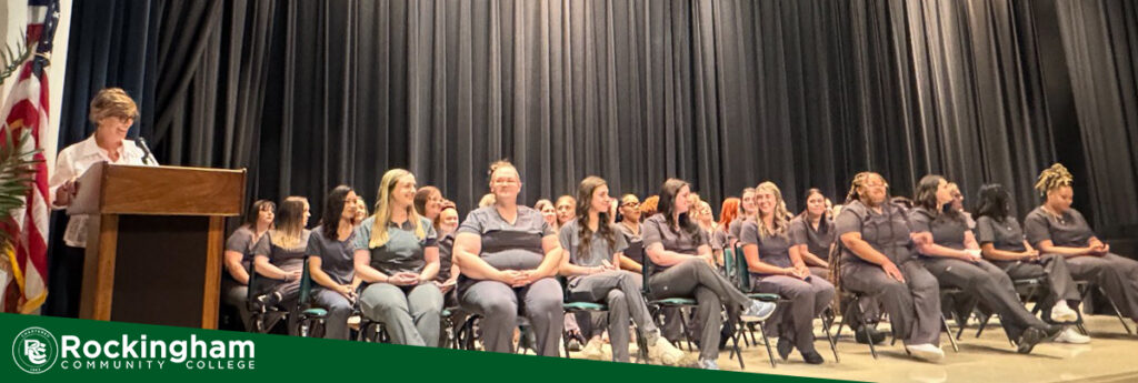 A woman speaks at a podium, students in black scrubs sit in chairs on a stage, and a green stripe in the bottom left incudes a logo for Rockingham Community College.