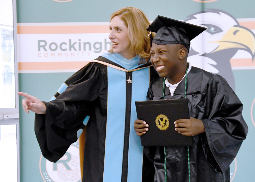 A graduate in a cap and gown holding his diploma grins ear to ear as he looks in the direction that a woman is pointing.