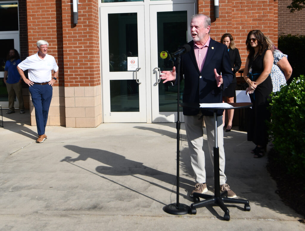 A man talks at a podium, with several people gathered behind him