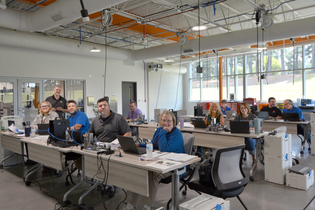 Adults in an industrial technologies lab sit at tables with laptops, as an instructor stands in the background.