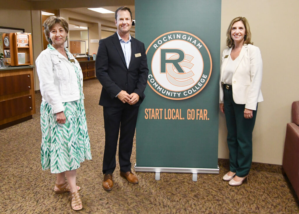 Two women and a man stand by a sign showing the Rockingham Community College logo.