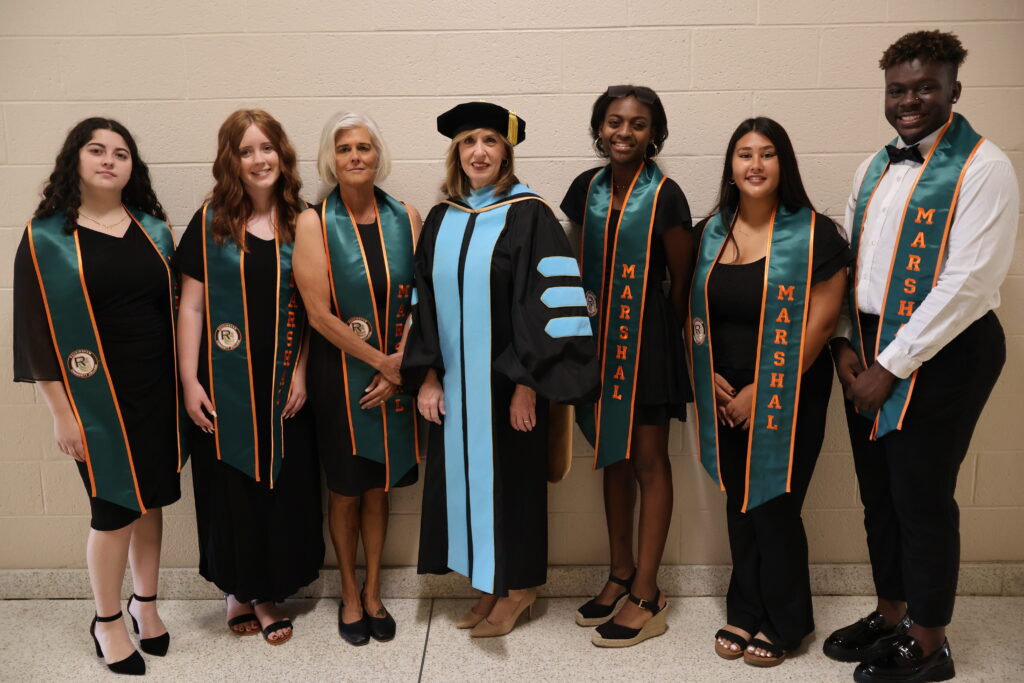 A group of women and a man wearing stoles that say marshal, are joined by a woman in academic regalia.
