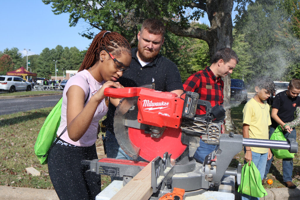 A young female student uses a miter saw under the watchful eye of an adult, as an adult and two students watch something else in the background.