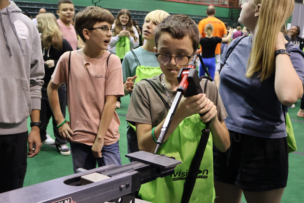 A young boy uses a virtual welder, and man other young students are in the background enjoying a career expo.