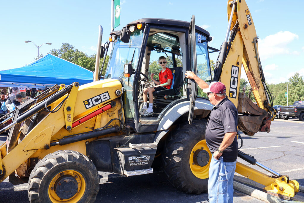 A young boy sits in a backhoe loader, as a man stands at the open door talking with him.