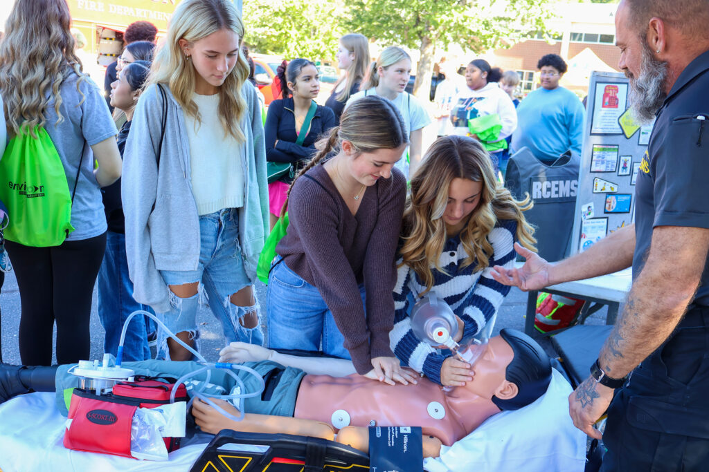A teenage girl gives CPR to a medical manikin while another gives it oxygen, as another watches and a man gives them directions. A crowd of teenagers is in the background.