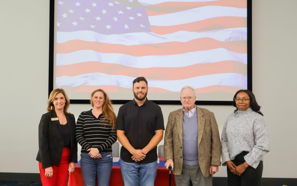 Three women and two men smile in front of a giant American flag on a monitor in the background.