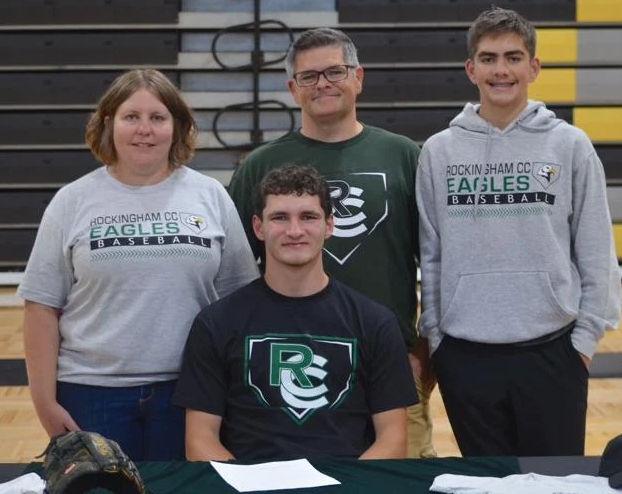 A teenager sits at a table and another teen and a couple stands behind him.