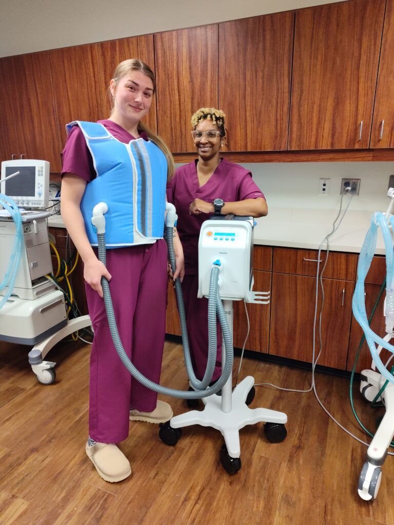 A lady in scrubs stands by a machine while another wears a respiratory therapy vest.