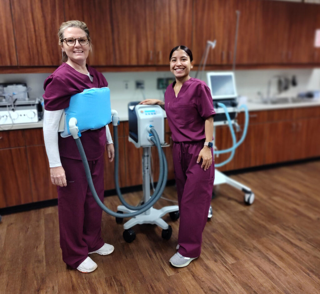 A lady in scrubs stands by a machine while another wears a respiratory therapy band around her chest.