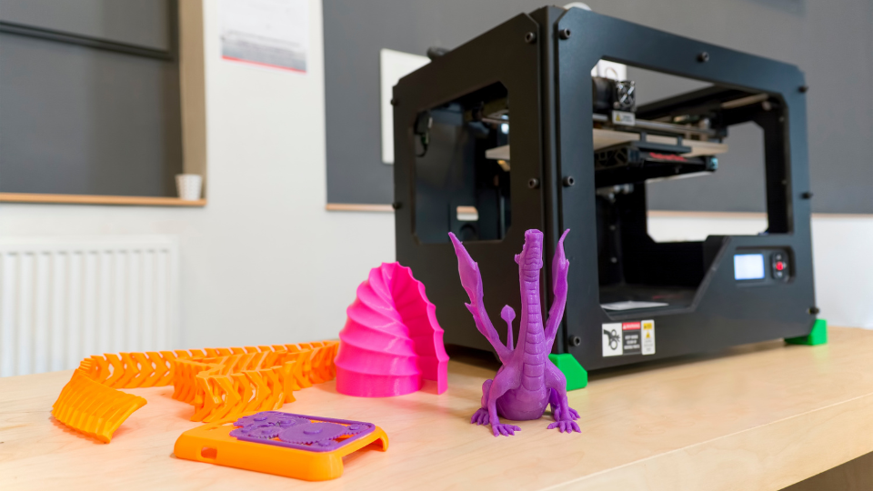 A 3D printer sitting on a table with items printed in 3D, a purple dragon, half a pink conch shell, an orange spine-looking long piece, and what looks like an orange phone case with purple gears on the back.