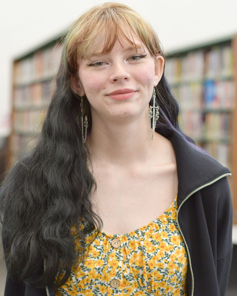 A girl in a yellow floral dress and black zippered sweatshirt smiles, with blurred out library stacks in the background.