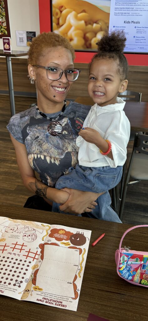 A mother and toddler smile, sitting at a table in a restaurant.
