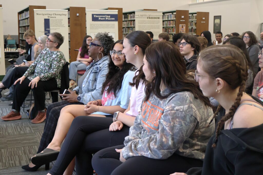 A full audience sitting in rows of chairs in a library.