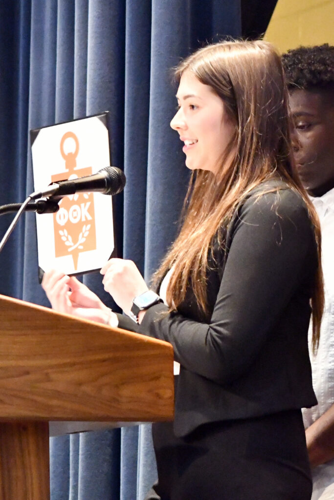 PTK Secretary Layla Ross explains the symbolism of the PTK badge, which she is holding up at the podium.