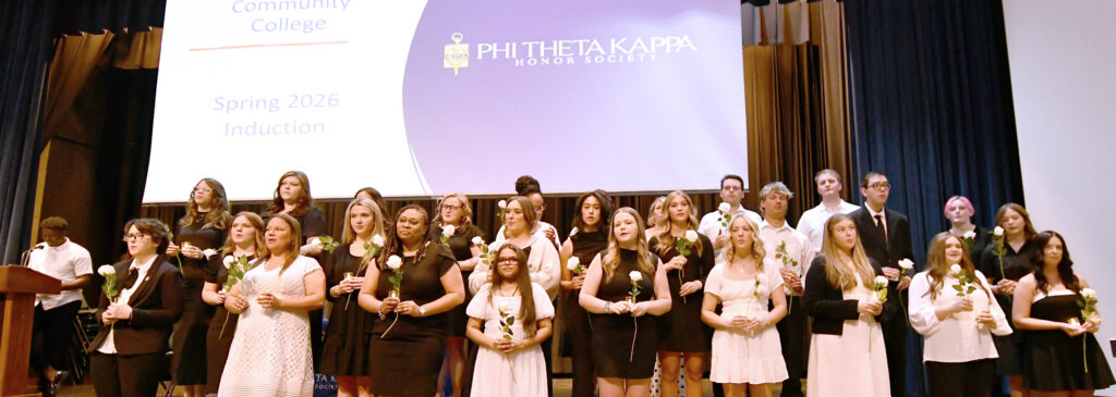 A group of more than 30 students stand on a stage under a Phi Beta Kappa sign, holding candles and white roses, as they take their oaths.