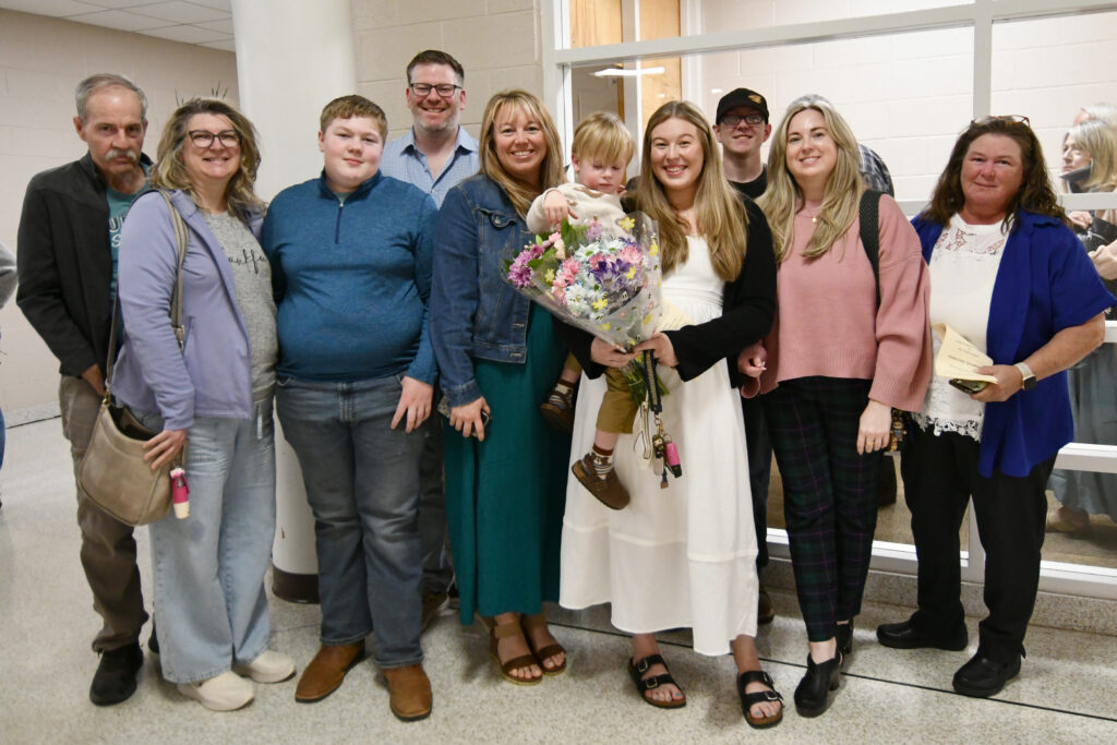 A student holds a large bouquet of flowers and a young boy, and stands in the middle of eight other family members.