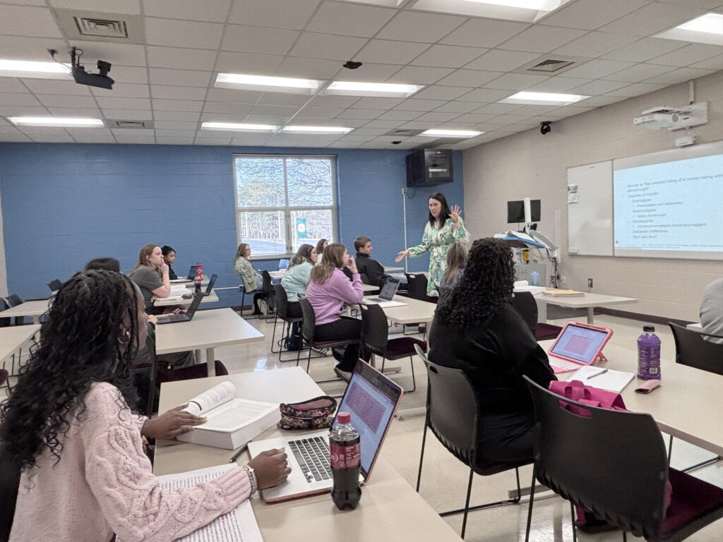 An instructor teaches a classroom of students, with notes being projected on a whiteboard, and cameras on the ceiling that enable her to be connected with students at home.