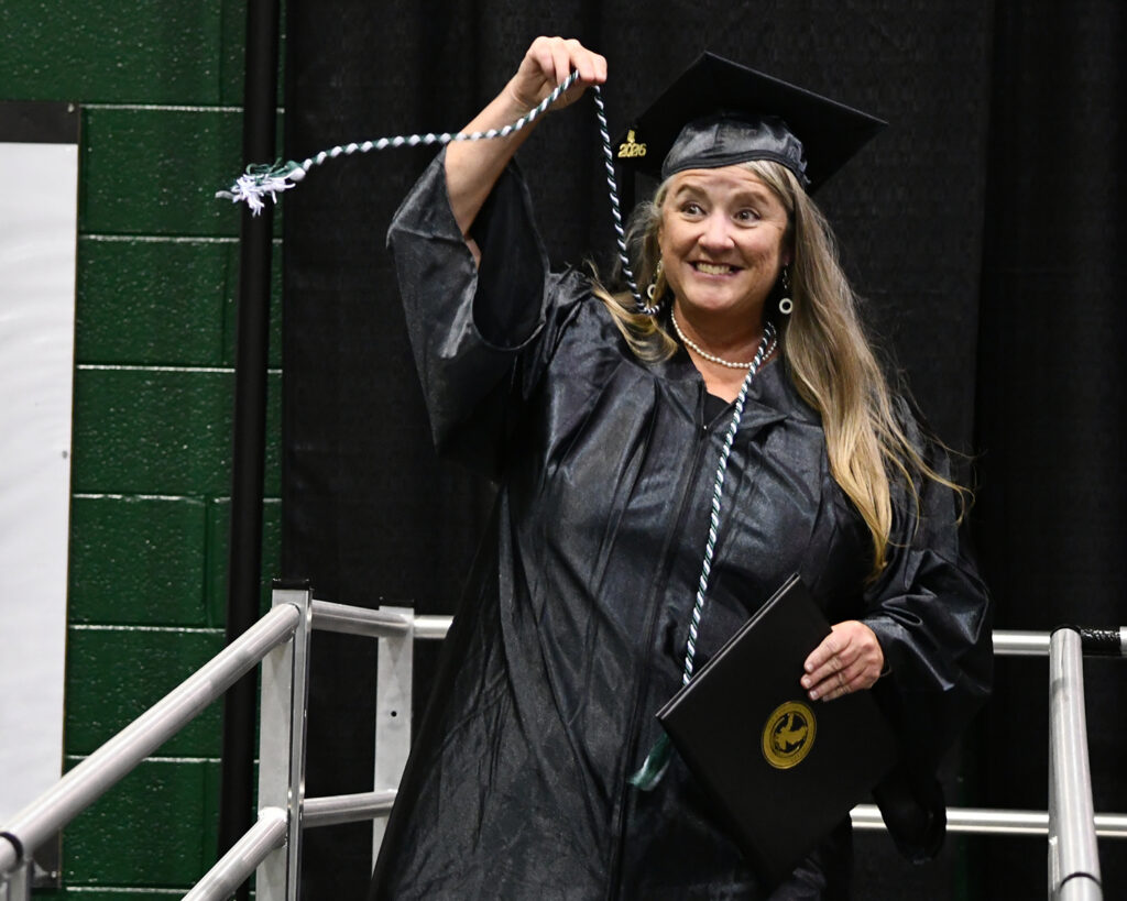 A graduate grins into the audience and holds up an honor cord.