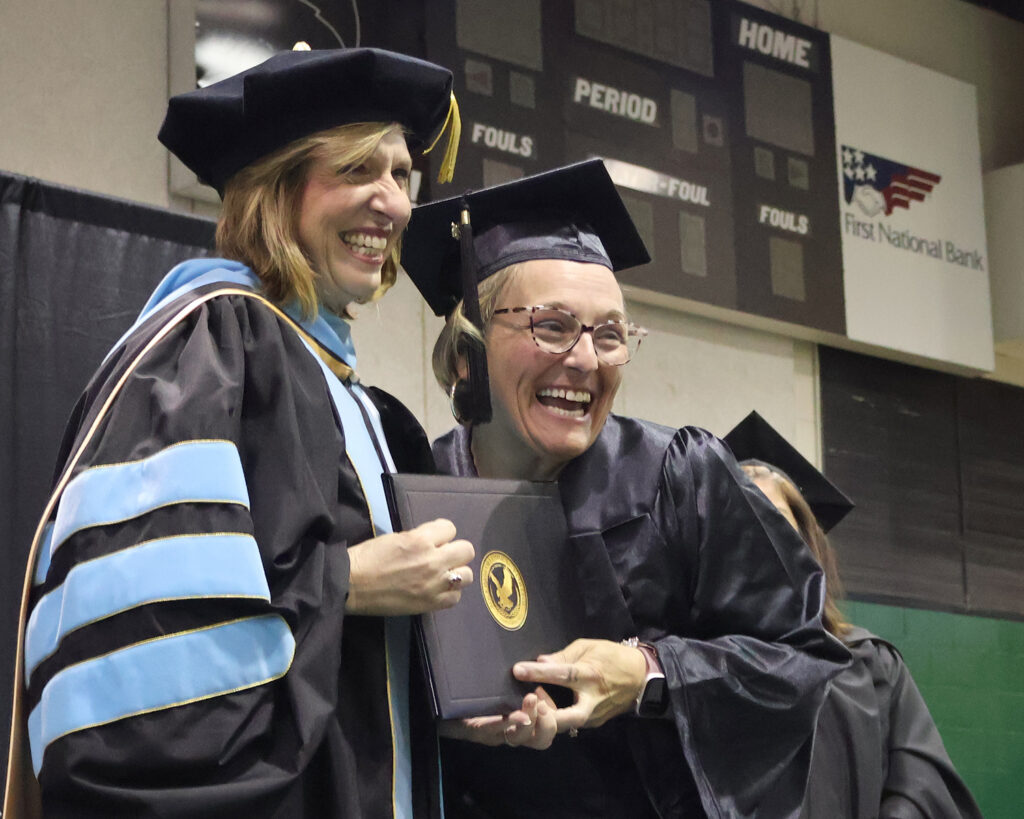 The college president and an older female graduate laugh on stage as the graduate accepts her diploma.