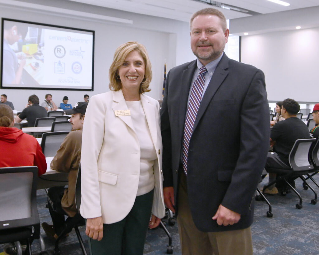 A man and a woman smile, with people sitting at tables behind them in a large room.